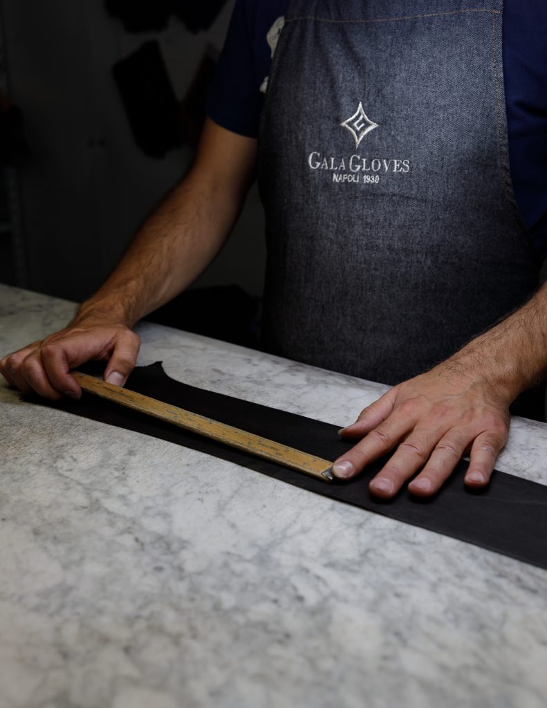 Artisan measuring black leather with wooden ruler wearing Gala Gloves Napoli 1930 branded apron - precision Italian craftsmanship at work