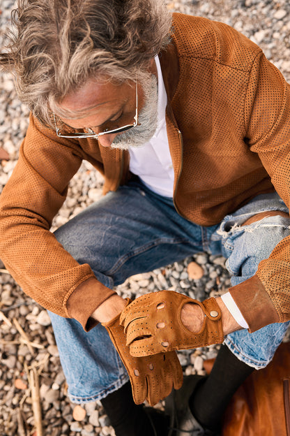 A man wearing brown perforated leather driving gloves adjusts them while seated on gravel, showcasing Italian craftsmanship.