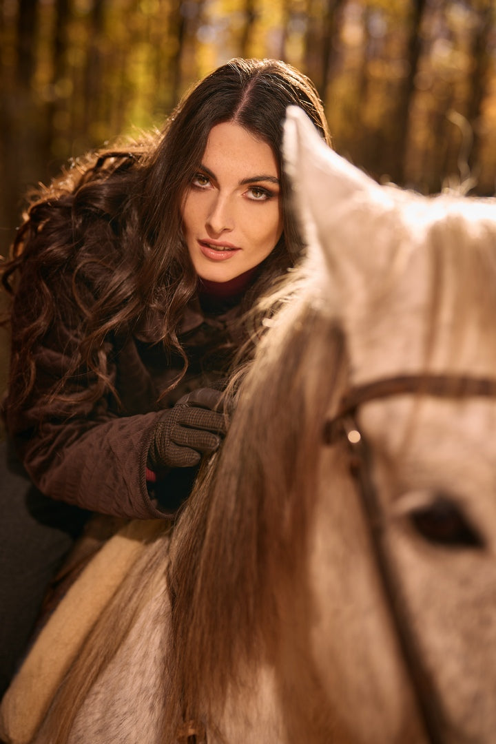 A woman in a brown jacket gently holds a white horse, showcasing elegant brown leather gloves in a serene forest setting.