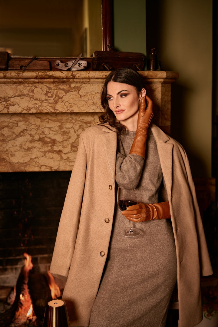 Elegant woman in cognac nappa leather gloves, holding a wine glass, styled with a beige coat by a cozy fireplace.
